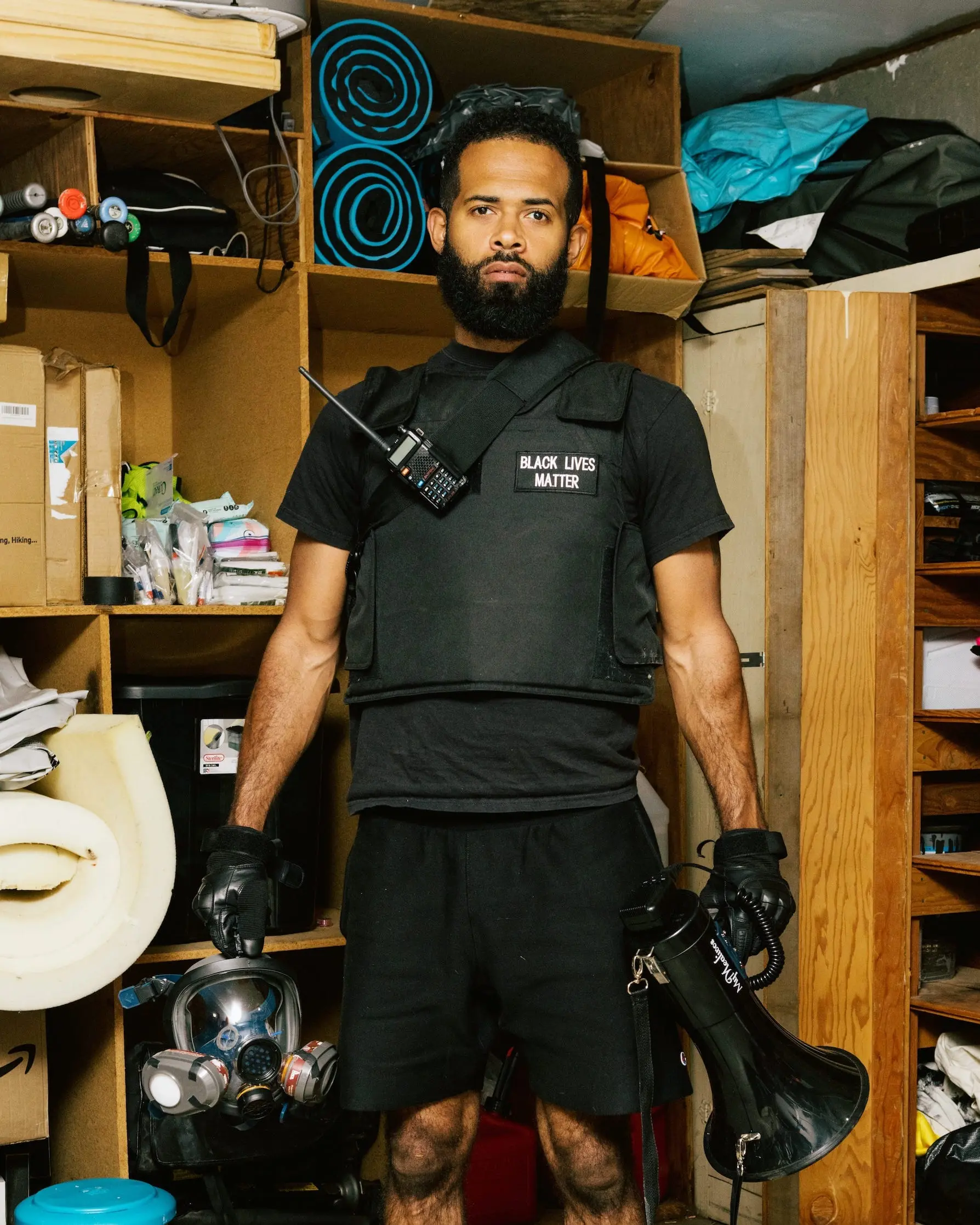 A man stands in black, protective gear, while holding a bullhorn and a gas mask in front of some shelves.